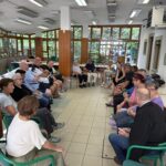 Slika prikazuje naše člane in njihove spremljevalce kako sedijo na stolih v krogu in poslušajo predavanje.