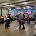 Na sliki je velika, polna dvorana, v kateri za okroglimi mizami sedi veliko članov. V ospredju in osrednjem delu prostora je plesišče, kjer se ob glasbi vrtijo plesni pari. Vzdušje je živahno in praznično, prostor pa deluje topel in družaben.