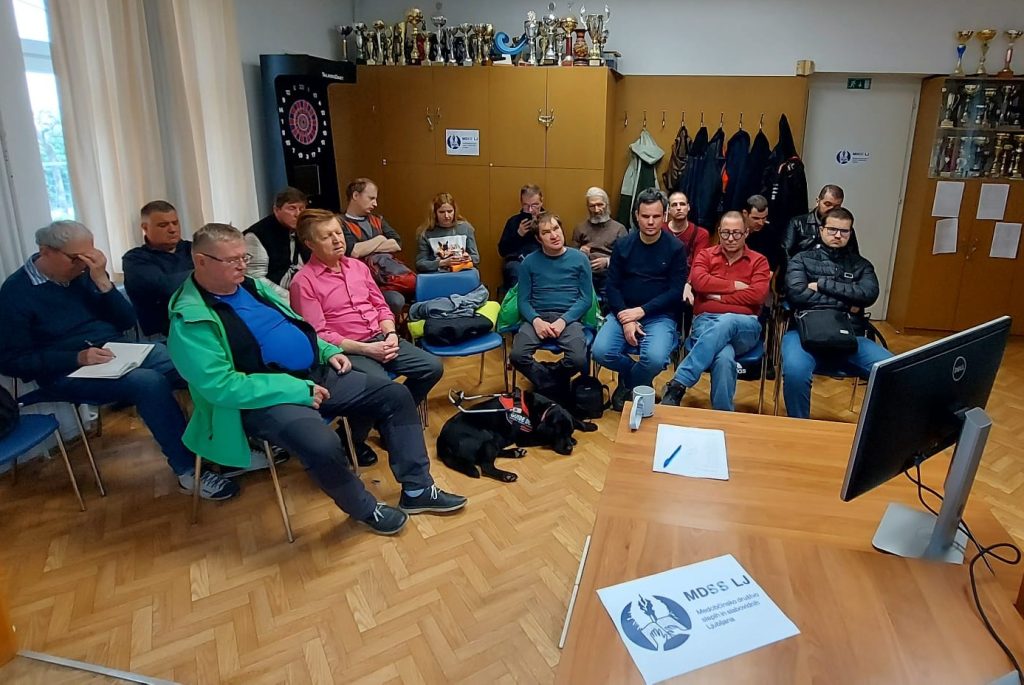 Videkonferenčno protidoping izobraževanje športnikov MDSS Ljubljana. 2.4.2026 v društvenih prostorih. Na fotografiji slušatelji izobraževanja, posedeni v dveh polkrožnih vrstah, med poslušanjem predavatelja (na sredini fotografije tudi pes vodnik).